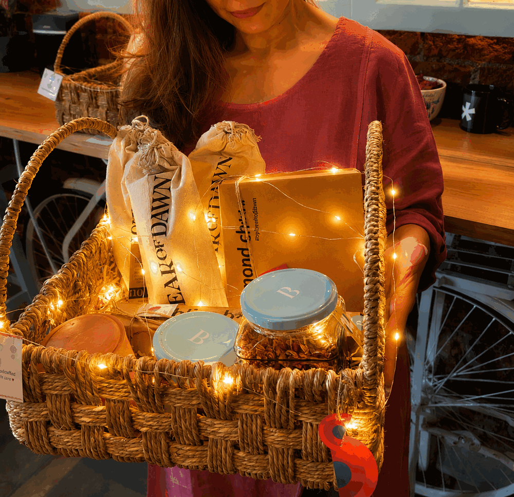 The large jute basket held in both hands, holding cloth bags, a box of almond cheese and lidded jars, lit with fairy lights.